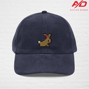 K&ouml;nnte beinhalten: Marineblaue Cord-Baseballkappe mit einem pixeligen, braunen Hasen-Design, das auf der Vorderseite gestickt ist. Die Kappe hat einen gebogenen Schirm und einen Knopf oben. Der Markenname "ATELIER DECODE" ist oben rechts aufgedruckt.