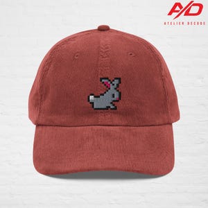K&ouml;nnte beinhalten: Rostfarbene Cord-Baseballkappe mit einem pixeligen grauen Hasen mit rosa Ohren. Die Kappe hat einen gebogenen Schirm und einen Knopf oben. Das Logo "A/D ATELIER DECODE" ist aufgedruckt.
