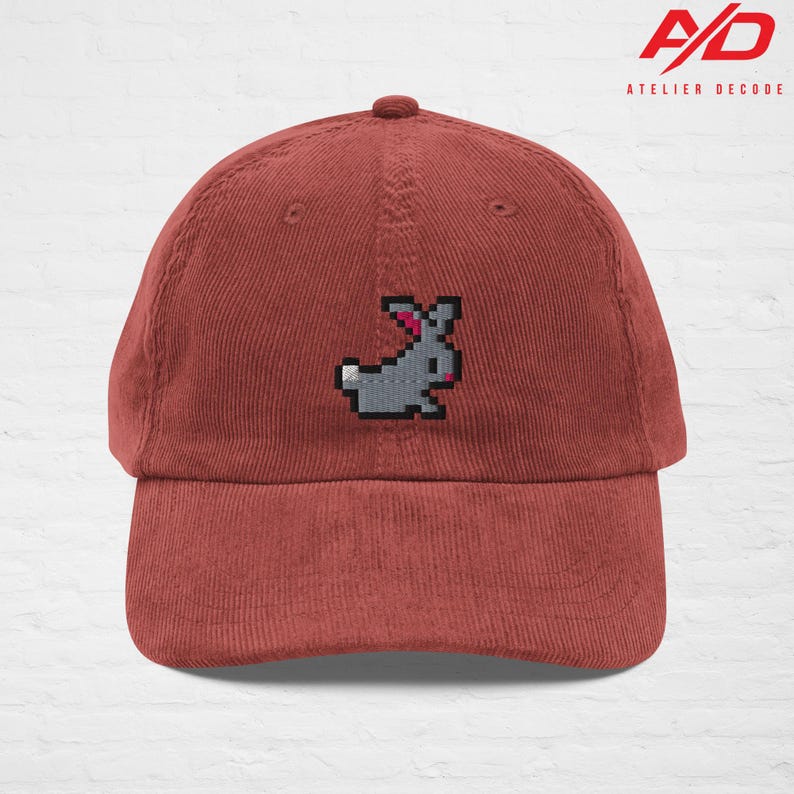 K&ouml;nnte beinhalten: Rostfarbene Cord-Baseballkappe mit einem pixeligen grauen Hasen mit rosa Ohren. Die Kappe hat einen gebogenen Schirm und einen Knopf oben. Das Logo "A/D ATELIER DECODE" ist aufgedruckt.