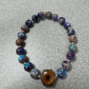 May include: A beaded bracelet featuring round, multi-colored stone beads in shades of blue, purple, and brown, accented with small silver-toned spacers. The focal point is a hexagonal charm with a central amber-colored eye graphic design.
