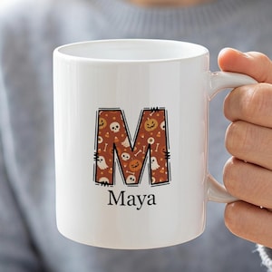 Puede incluir: Una taza de cerámica blanca con una letra M grande rellena de un patrón de calabazas naranjas, fantasmas blancos, calaveras y huesos sobre un fondo oscuro. Debajo de la letra M, el nombre Maya está impreso en una fuente serif negra.