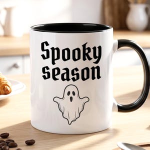 Könnte beinhalten: Ein weißer Keramikbecher mit schwarzem Innenraum und schwarzem Henkel. Auf der Vorderseite befindet sich der schwarze Schriftzug "Spooky Season" in einer stilisierten Schrift über einer einfachen schwarzen Strichzeichnung eines Geistes.