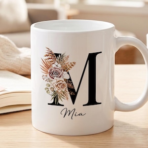 Pode incluir: Uma caneca de cerâmica branca com uma letra M maiúscula grande em preto, decorada com um arranjo floral de flores secas, erva-das-pampas e rosas. O nome Mia está impresso numa fonte cursiva preta abaixo da letra M.