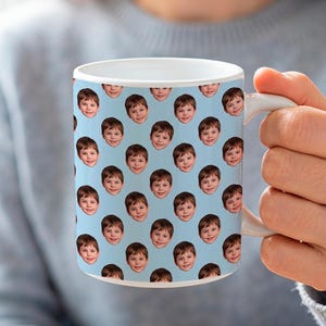 Puede incluir: Una taza de cerámica blanca con un patrón repetitivo del rostro de un niño pequeño impreso en la superficie exterior. El fondo de la taza es de color azul claro sólido y cuenta con un asa curva en el lado derecho.