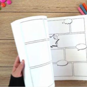 May include: A blank comic book with several panels and speech bubbles is being held open. The pages are white with black outlines. Colored markers, magnets, and a Rubik's cube are scattered on a wooden surface.