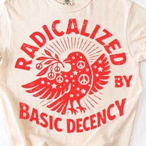 May include: Cream-colored t-shirt with the words "RADICALIZED BY BASIC DECENCY" in red, surrounding a red dove carrying an olive branch. The dove has peace signs and stars.