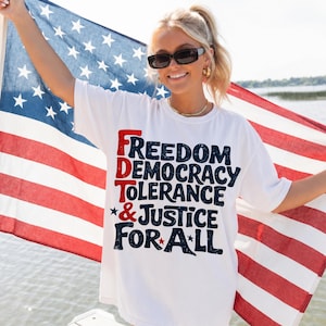 Può includere: Maglietta bianca con le parole "Freedom, Democracy, Tolerance & Justice For All" in lettere rosse e blu. La persona tiene in mano una bandiera americana a strisce rosse e bianche e stelle blu.