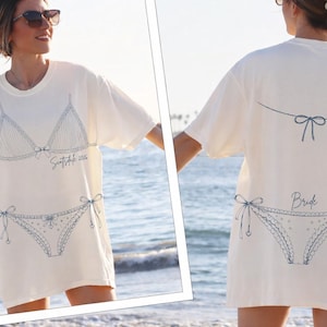 Puede incluir: Camiseta blanquecina con un diseño de bikini azul en la parte delantera y trasera. La parte delantera presenta un sujetador y una braguita de bikini con el texto "Suntribe 2026". La parte trasera de la camiseta dice "Bride".
