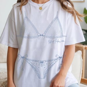 Könnte beinhalten: Weißes T-Shirt mit einer blauen Strichzeichnung eines Dessous-Sets. Das Design umfasst einen BH mit vertikalen Streifen und Spitzenbesatz sowie einen Slip mit Schleifen- und Herzdetails. Der Text "Isle of Palms 2026" ist aufgedruckt.