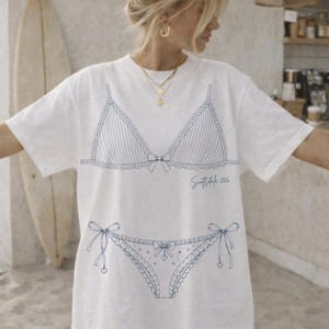 May include: White t-shirt with a blue line drawing of a lingerie set. The design includes a bra with vertical stripes and a bow, and matching briefs. The text "Scottsdale 2026" is written in cursive below the bra.