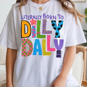 May include: White t-shirt with the phrase "Literally Born To Dilly Dally" in colorful, playful lettering. The design features a flower, polka dots, and various patterns within the letters, creating a fun and vibrant look. The shirt is casual and comfortable.