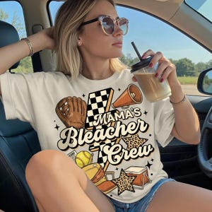Puede incluir: Camiseta color crema con el texto "Mama's Bleacher Crew" en un diseño vintage. El gráfico incluye un guante de béisbol, un megáfono, estrellas y una bandera a cuadros. La camiseta es de estilo casual y deportivo.
