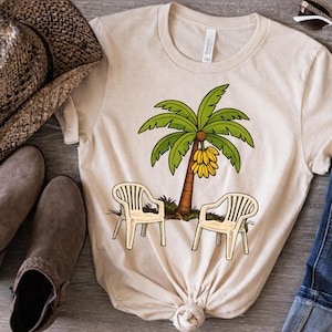 Puede incluir: Camiseta color crema con un diseño de palmera de dibujos animados con hojas verdes y plátanos. Dos sillas de jardín de plástico blanco frente al árbol. Prenda de vestir informal.