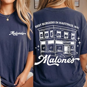 May include: Navy blue t-shirt with the text "Malone's" on the front and a detailed white illustration of a restaurant with the text "Best Burgers in Hastings, MA!" on the back. The shirt is a casual style.