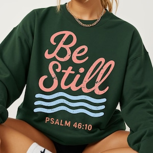 May include: A dark forest green crewneck sweatshirt featuring the text "Be Still" in a distressed pink script font, followed by three light blue wavy lines and the text "PSALM 46:10" in a simple, distressed pink sans-serif font.