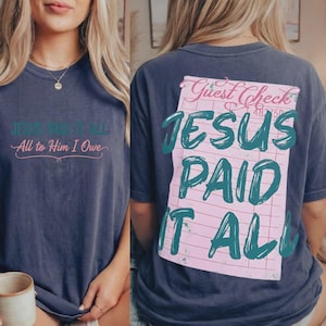 May include: Navy blue t-shirt featuring the text "JESUS PAID IT ALL" on the back, printed on a pink guest check design. The front of the shirt has the text "JESUS PAID IT ALL, All to Him I Owe" in teal and pink.