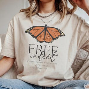 Può includere: T-shirt beige con un disegno di farfalla arancione e nera. Il testo "FREE indeed" è stampato sotto, insieme a un versetto biblico. La maglietta è in materiale morbido. Un top casual.