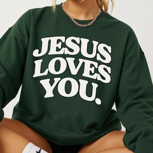May include: Dark green sweatshirt with the text "JESUS LOVES YOU" in large, white, bold letters. The sweatshirt has a crew neck and long sleeves. A gold chain necklace is visible at the neckline. The overall style is casual and comfortable.