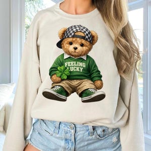 Può includere: Una felpa color crema con una grafica di un orsacchiotto che indossa un maglione verde con la scritta "FEELING LUCKY" e tiene in mano un quadrifoglio. L'orso indossa anche un cappello a scacchi, pantaloni color cachi e scarpe da ginnastica verdi.