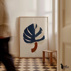 May include: Large framed wall art featuring a stylized monstera leaf design. The leaf is split into sections of navy blue and off-white, with gold accents. The stem is a rust color. The artwork is in a room with a small wooden side table.