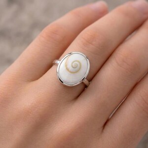 Può includere: Anello ovale con un design a conchiglia bianca e un motivo a spirale centrale. L'anello ha una fascia argentata e un'incastonatura a castone. L'anello è mostrato su un dito.