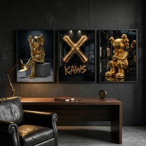 May include: Three framed art prints showcasing gold KAWS figures and a gold balloon 'X' with the word 'KAWS'. The prints are above a dark wood desk with a black leather chair and a gold desk lamp.
