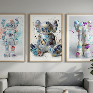 May include: Three framed art prints featuring colorful, graffiti-style bear figures. Each bear is white with black cross-shaped eyes and various colorful accents. The backgrounds have paint splatter effects in shades of blue, pink, and orange.