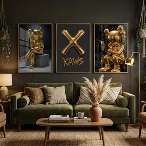 May include: Three framed art prints. The prints feature gold sculptures and a gold 'X' with the word 'KAWS'. They are displayed above a green sofa with decorative cushions. The room has a dark, moody aesthetic.