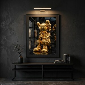 May include: A framed artwork showing a gold sculpture of a seated figure with a unique head shape. The artwork is displayed on a dark wall above a black console table. A black vase with a decorative branch and books are also visible.