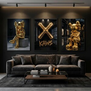 May include: Three framed art prints. Two feature gold sculptures, one seated on a grey block. The third shows a gold balloon 'X' with 'KAWS' text. The background is dark, with a modern interior setting.