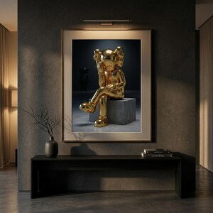 May include: A framed artwork showing a gold sculpture of a seated figure with a unique head design. The sculpture is on a grey cube against a dark backdrop. The artwork is displayed above a black console table.