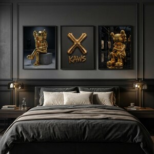 May include: A bedroom scene with three framed artworks. The artworks feature gold sculptures and a gold "X" with the word "KAWS". The bed has a dark grey duvet and pillows. Two gold lamps sit on the bedside tables.