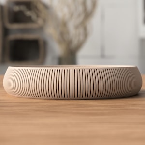 Könnte beinhalten: Eine niedrige, runde, dekorative Schale in hellem Beige mit vertikalen Rillen. Die Schale steht auf einer Holzoberfläche. Der Hintergrund ist unscharf und zeigt ein neutral getöntes Interieur mit dekorativen Elementen. Das Design der Schale deutet auf eine moderne Ästhetik hin.