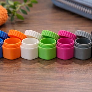 Könnte beinhalten: Eine Sammlung kleiner, farbenfroher Plastikdosen mit passenden Schraubdeckeln. Die Dosen sind in verschiedenen Farben erhältlich, darunter Blau, Orange, Weiß, Grün, Pink, Grau und Schwarz. Die Deckel haben ein geriffeltes Design.