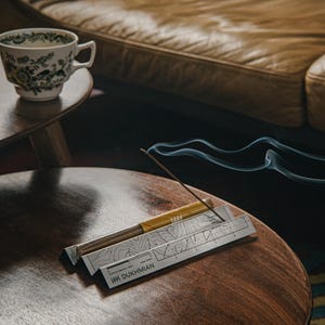 Könnte beinhalten: Eine Tasse und Untertasse mit floralem Muster stehen auf einem Holztisch. Ein Räucherstäbchen brennt, mit aufsteigendem Rauch. Der Räucherstäbchenhalter hat ein Kartendesign und den Text "HH DUKHMIAN". Im Hintergrund ist ein Ledersofa zu sehen.