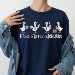 Puede incluir: Una camiseta azul marino con un gráfico de cuatro patos blancos realizando tareas de oficina y el texto "Para Normal Activities". El primer pato usa un portátil, el segundo sostiene un portapapeles y un megáfono, el tercero lleva papeles y una taza de café, y el cuarto organiza una estantería.