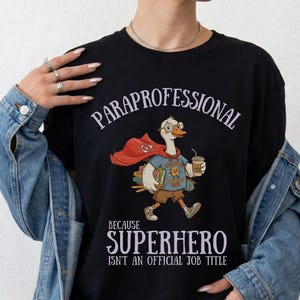 Puede incluir: Una camiseta negra con el gráfico de un ganso con gafas, una capa roja y cargando libros y un vaso de café. El texto dice PARAPROFESSIONAL BECAUSE SUPERHERO ISN'T AN OFFICIAL JOB TITLE. El ganso viste una camiseta azul y pantalones cortos marrones.