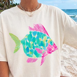 Könnte beinhalten: Cremefarbenes T-Shirt mit einem farbenfrohen Aquarell-Fisch-Design. Der Fisch hat einen türkisfarbenen Körper mit grünen und gelben Akzenten und rosa Flossen. Das Shirt hat eine lockere Passform, ideal für den Strand.