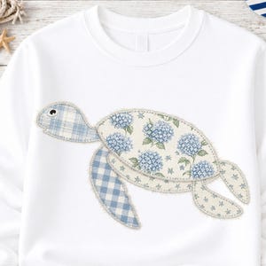 May include: White sweatshirt featuring a whimsical turtle design. The turtle is crafted from patches of blue gingham and floral fabric, with a light blue and white hydrangea pattern. The design has a playful, handcrafted aesthetic.