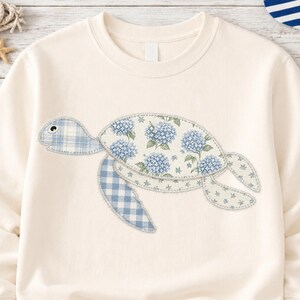 May include: Cream-colored sweatshirt featuring a sea turtle design. The turtle is made of patches of blue floral and gingham fabric. The design is centered on the front of the sweatshirt, creating a nautical theme.