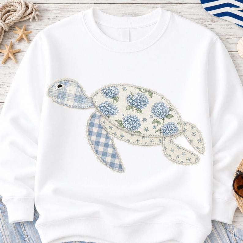 May include: White sweatshirt featuring a whimsical turtle design. The turtle is crafted from patches of blue gingham and floral fabric, with a light blue and white hydrangea pattern. The design has a playful, handcrafted aesthetic.