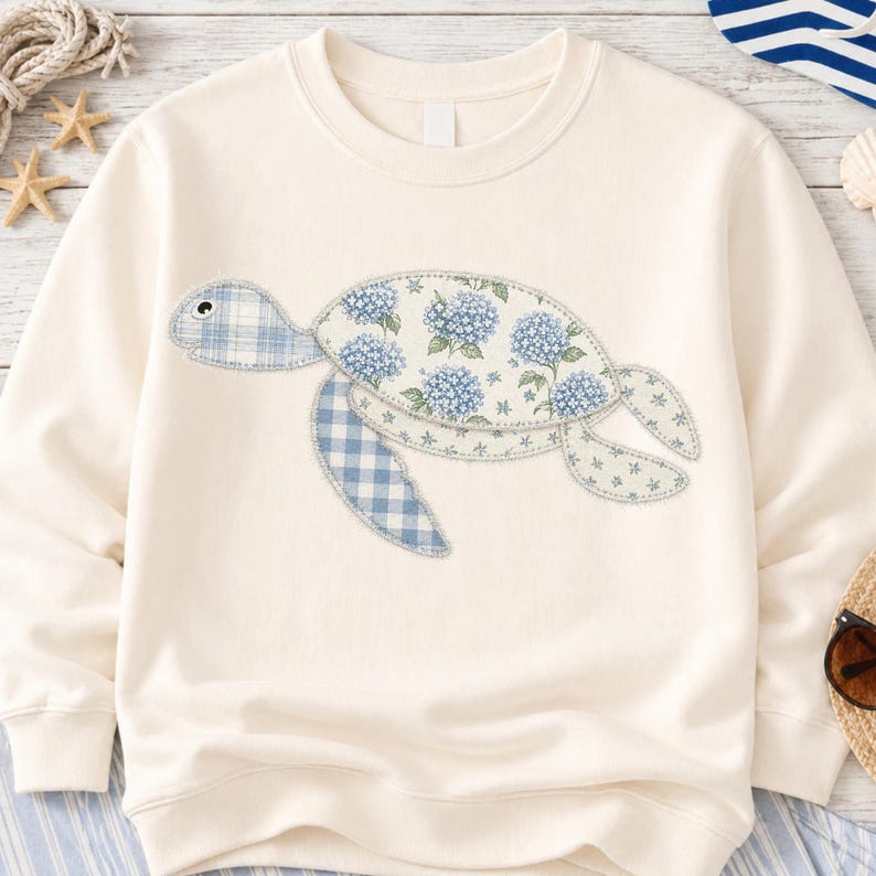 May include: Cream-colored sweatshirt featuring a sea turtle design. The turtle is made of patches of blue floral and gingham fabric. The design is centered on the front of the sweatshirt, creating a nautical theme.