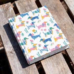 May include: A hardback book with a white cover featuring a repeating pattern of dachshund dogs in various colours including blue, yellow, and pink. The dogs are decorated with bows and floral accents in shades of green, pink, and yellow.