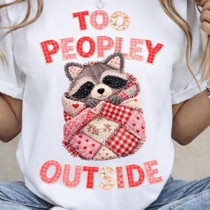May include: White t-shirt with the text "TOO PEOPLEY OUTSIDE" in red letters. A raccoon wrapped in a patchwork quilt is in the center. The quilt has pink hearts, floral patterns, and red gingham squares.