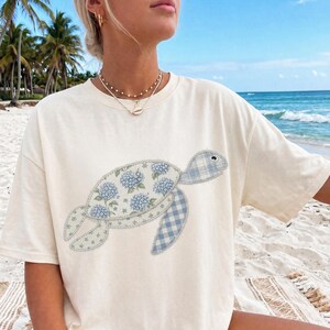 May include: Cream-colored t-shirt featuring a whimsical turtle design. The turtle's body is adorned with blue hydrangea flowers, while its flippers and head showcase a blue and white plaid pattern. The shirt is made of a soft material.