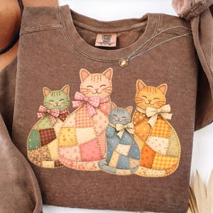 Puede incluir: Sudadera Comfort Colors marrón con un gráfico de cuatro gatos sonrientes. Cada gato tiene un diseño de patchwork y un lazo de diferentes colores: verde, rosa, azul y naranja. Se ve un delicado collar con un pequeño colgante.