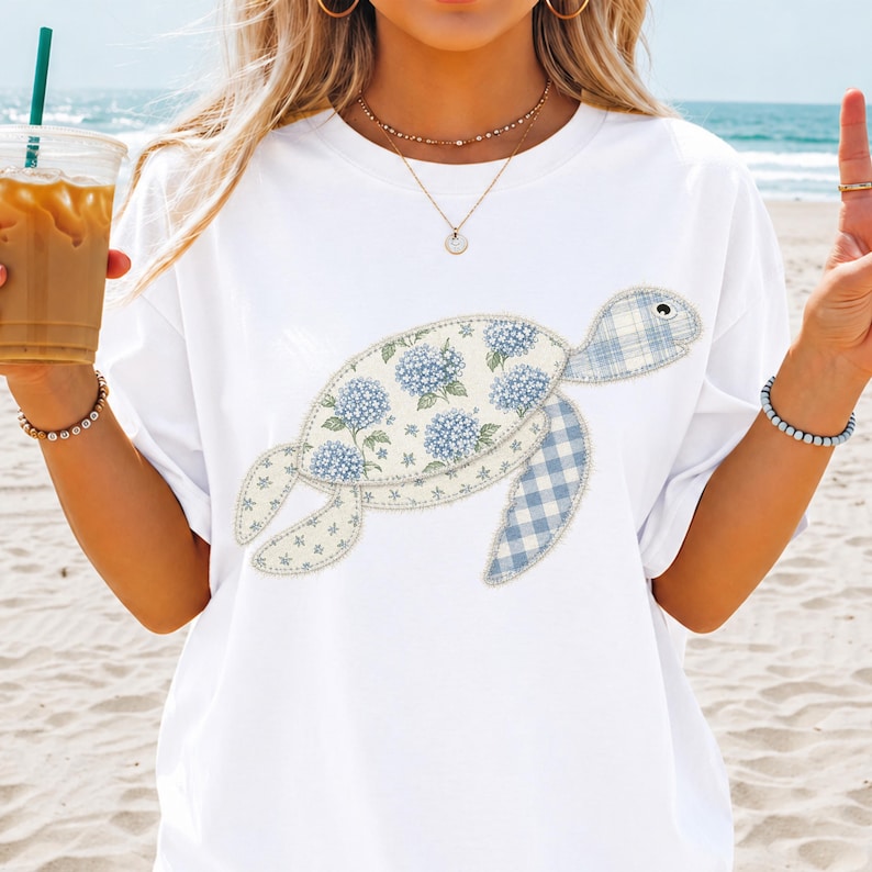 May include: White t-shirt featuring a turtle design. The turtle's shell is decorated with blue hydrangea flowers and a light blue gingham pattern. The turtle's flippers are also light blue gingham. The shirt is a casual style.