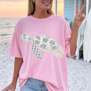 May include: A light pink t-shirt featuring a turtle design. The turtle has a floral pattern on its shell and blue gingham patterned flippers. The shirt is made of a soft material and is perfect for casual wear.