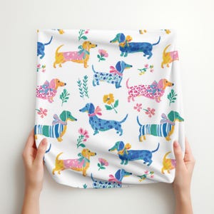 May include: A white fabric featuring a repeating pattern of dachshunds in various colours including blue, yellow, and pink. The dogs are illustrated with bows and wearing patterned clothing like stripes and spots, interspersed with small colourful floral motifs.