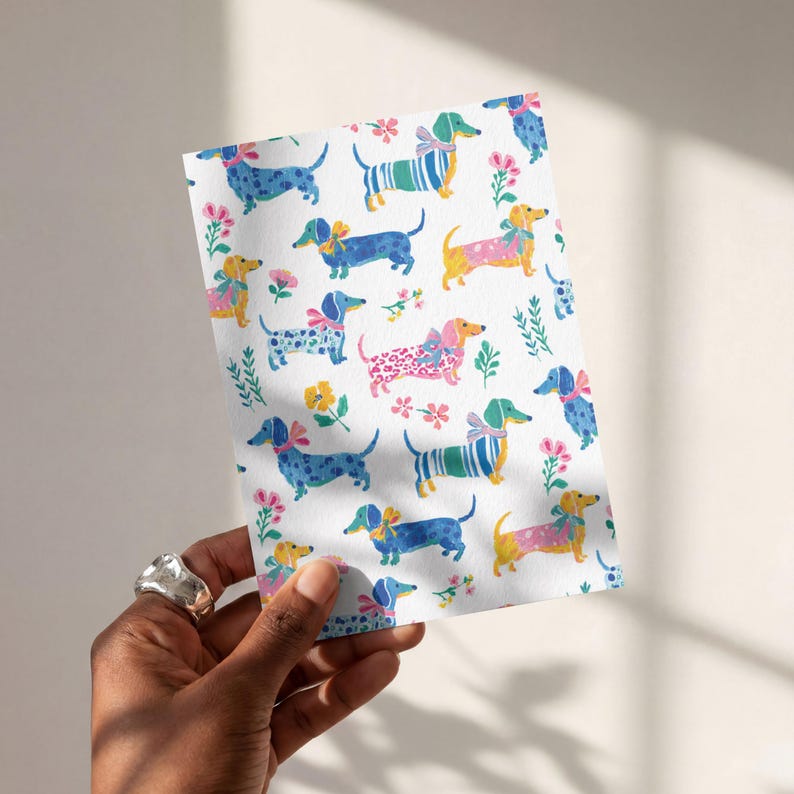 May include: A rectangular greeting card featuring a repeating pattern of dachshund dogs in various colours including blue, yellow, and pink. The dogs are decorated with bows and patterns, interspersed with small pink and yellow floral illustrations.